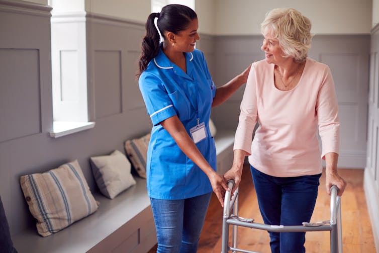 A woman in a nurse uniform assists an elderly woman using a walker.