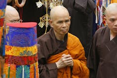 A bald man in an orange robe has his hands clasped in front of him.