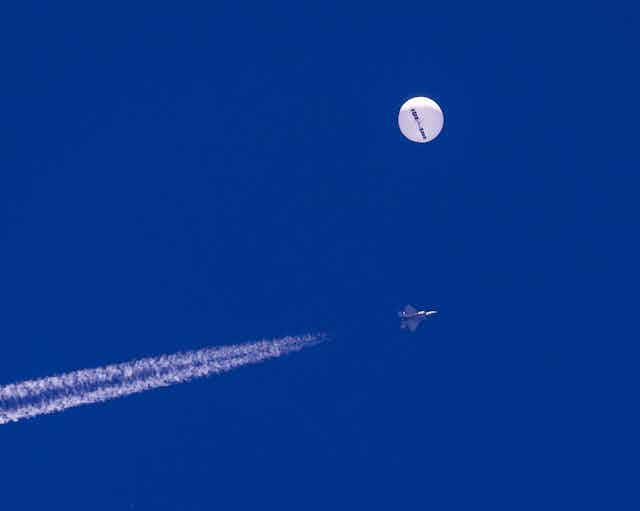 A large white balloon in the sky with a fighter jet and a contrail underneath it.
