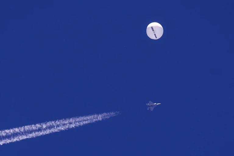 A large white balloon in the sky with a fighter jet and a contrail underneath it.