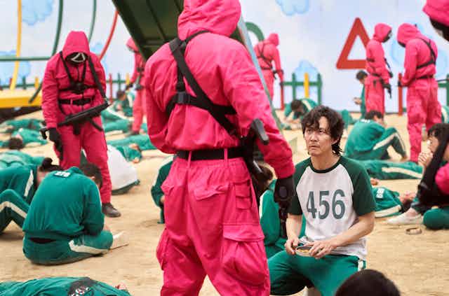 A man kneels in front a man in a red hazmat suit who is holding a gun.