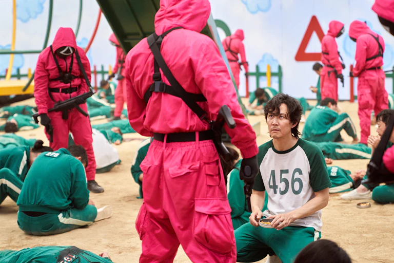 A man kneels in front a man in a red hazmat suit who is holding a gun.