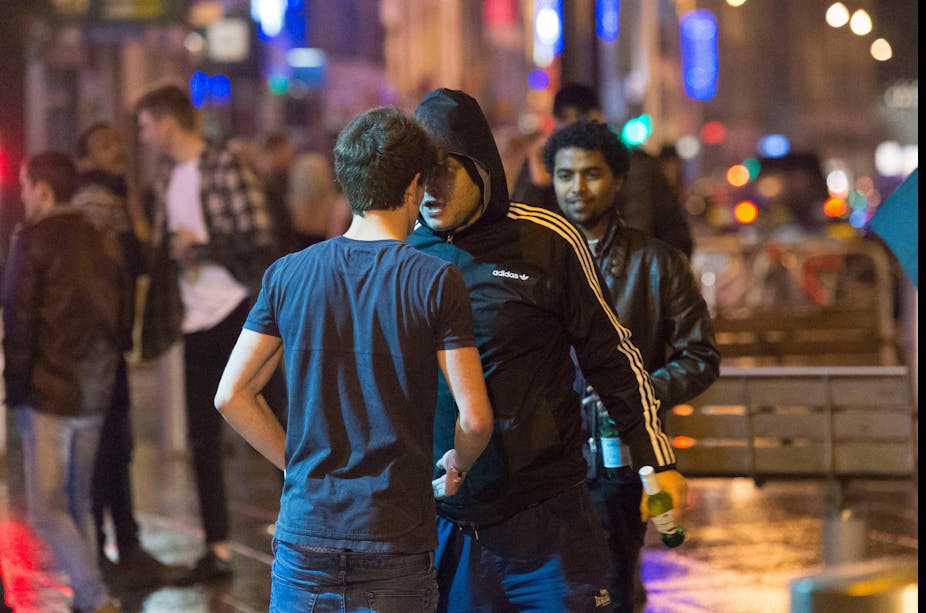 A man in a hooded Adidas top pushes his face into a man wearing a navy t-shirt. It is night time and there are people mingling in the background. 