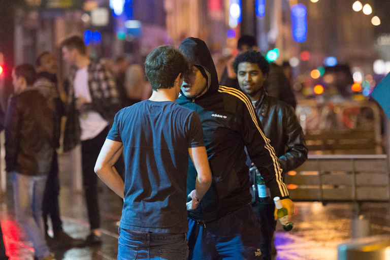A man in a hooded Adidas top pushes his face into a man wearing a navy t-shirt. It is night time and there are people mingling in the background.