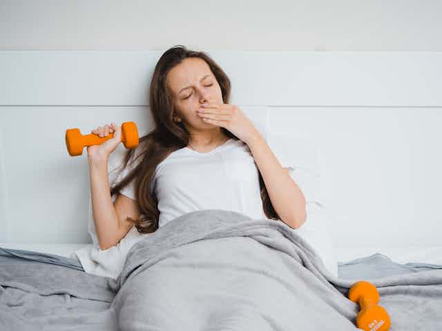 A yawning woman is tucked into bed, holding a dumbbell in one hand.