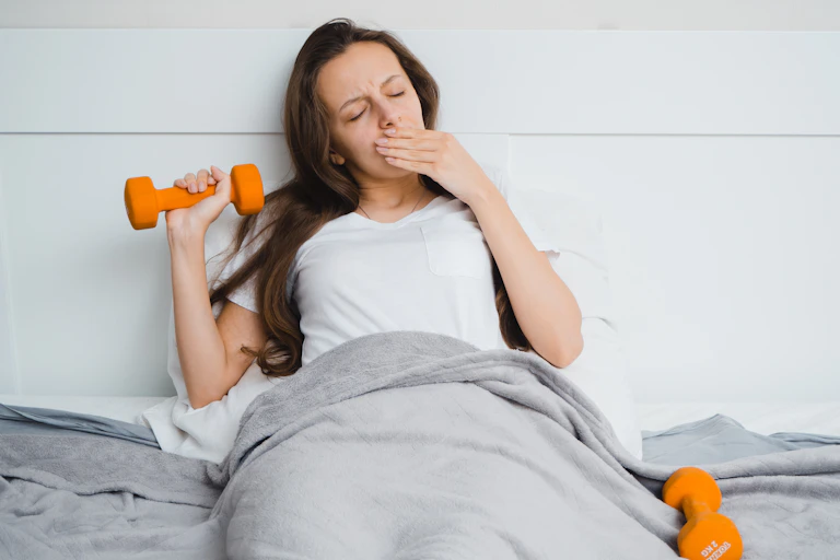 A yawning woman is tucked into bed, holding a dumbbell in one hand.