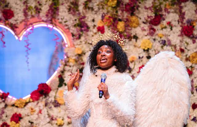 A young black woman sings into a microphone, wearing a crown and a dress and wings made of feathers. Behind her is a flower wall with a heart-shaped cut out.