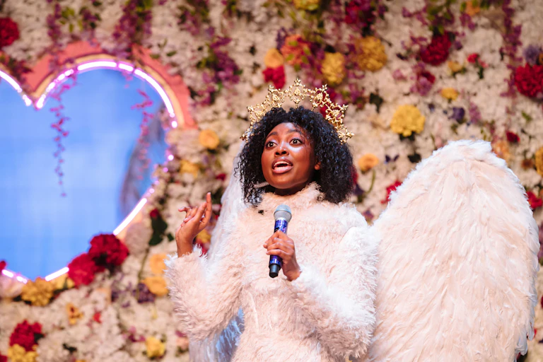 A young black woman sings into a microphone, wearing a crown and a dress and wings made of feathers. Behind her is a flower wall with a heart-shaped cut out.