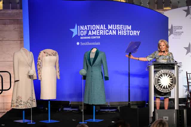 A middle aged white lady with blond hair stands at a podium and gestures towards mannequins with three outfits on them - 2 white, one blue. Behind her is a banner that says National Museum of American History.  