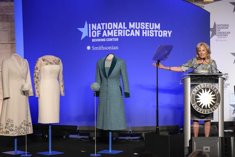 A middle aged white lady with blond hair stands at a podium and gestures towards mannequins with three outfits on them - 2 white, one blue. Behind her is a banner that says National Museum of American History.