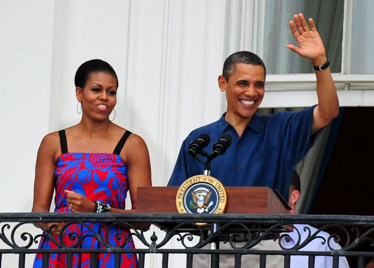 A Black middle aged man wearing a navy blue polo shirt waves and smiles on a balcony, next to a middle aged Black woman with a colorful red and blue patterned dress with thin black straps.