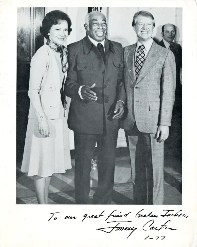 An elderly Black men is standing between a smiling white man and a white women.