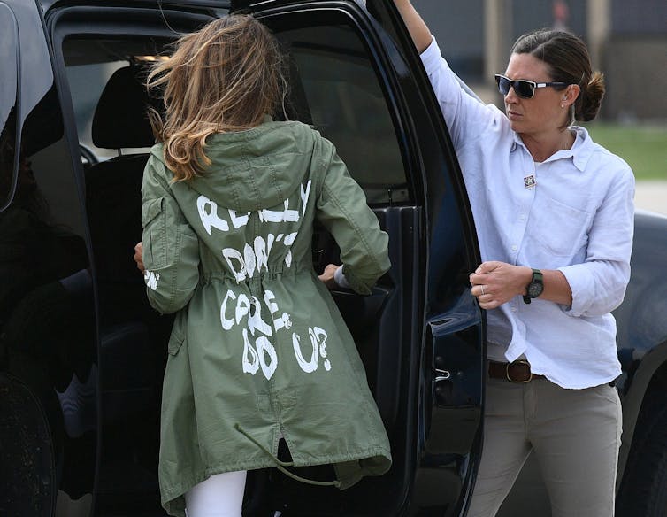 The back of a woman with long brown hair is shown getting into a black car. She wears an army green jacket with white writing. A woman wearing a white shirt holds open the car door for her.