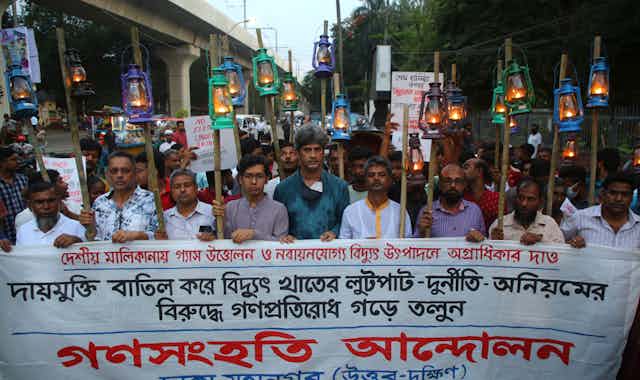 Protesters march down a street with a banner and kerosene lanterns on poles.