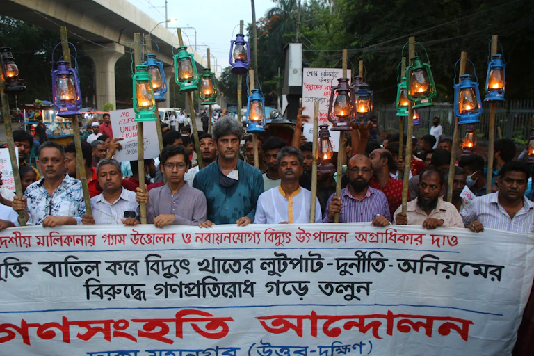 Protesters march down a street with a banner and kerosene lanterns on poles.