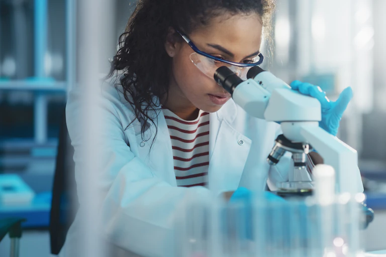 A woman wearing a white coat and safety glasses looking into a microscope