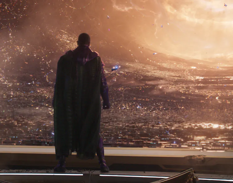 Kang wears a cape and facing away from the viewer, looks out over what seems to be a space city.