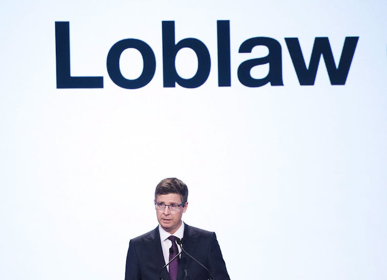 A man in a suit and tie wearing glasses speaking in front of a podium. Behind him is a massive screen that says 'Loblaw' on it