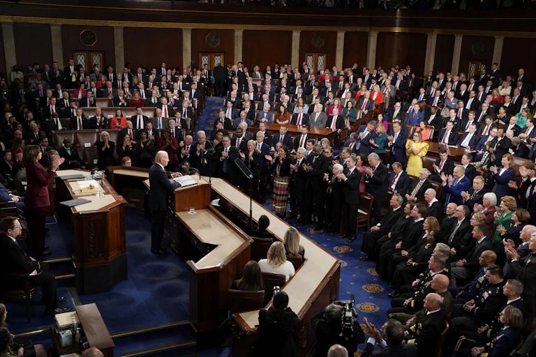 A white-haired man standing behind a large desk talking to a lot of people in a large hall.