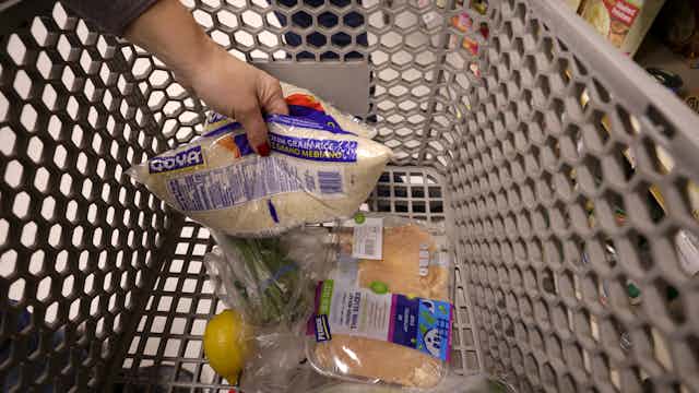 A grocery cart with a few items in it, shown as someone tosses in a bag of rice.