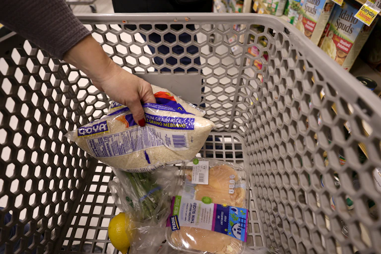 A grocery cart with a few items in it, shown as someone tosses in a bag of rice.
