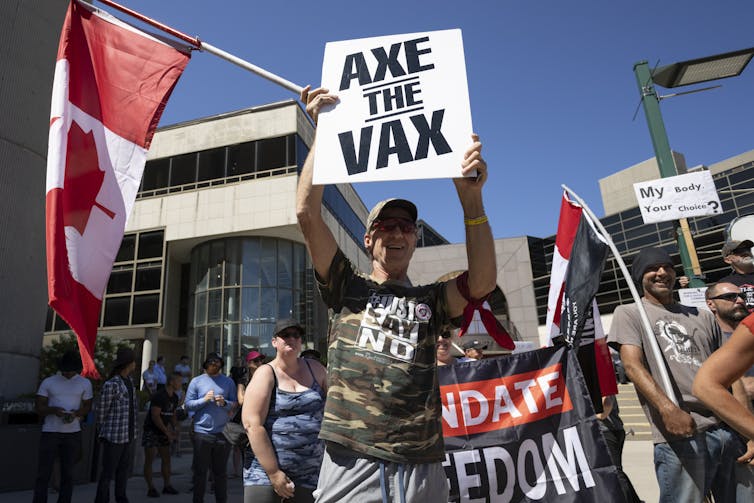 a man at a demonstration holds a Canadian flag and a sign that reads: Axe the vax.