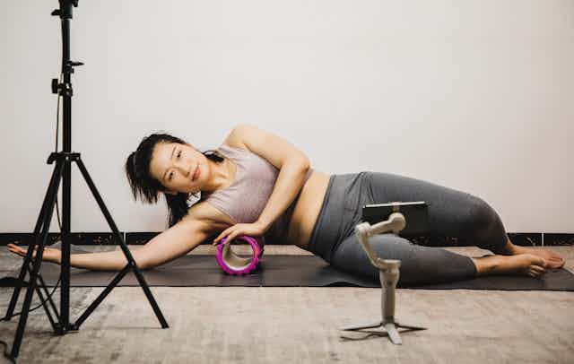 Woman stretching using foam roller while live streaming via phone on a tripod.