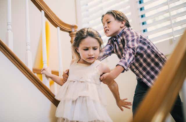 An angry boy grabs a fleeing girl's arm on a flight of stairs in a home.