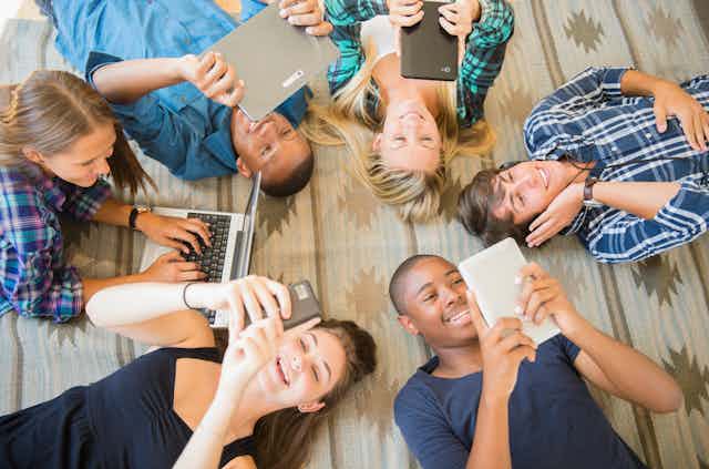 six teenagers lie on the floor looking at phones, tablets and a laptop