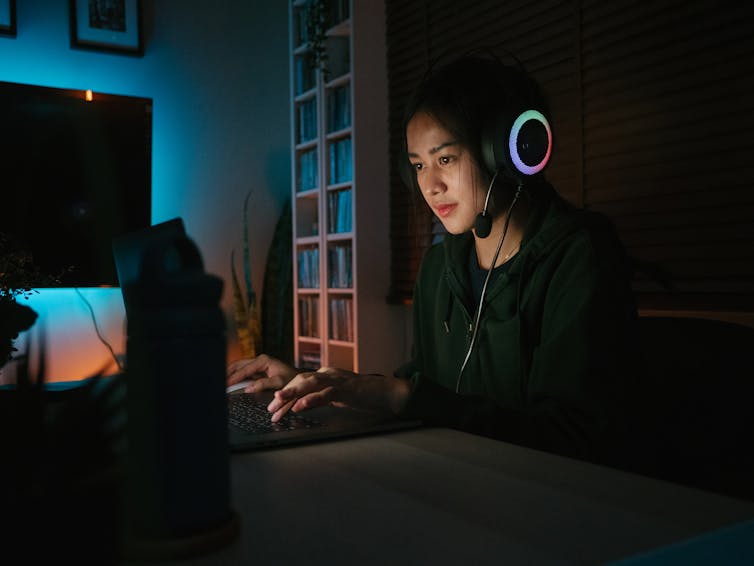 an Asian teenage girl wearing headphones in a dark room types on a laptop keyboard