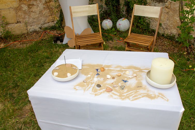 Sand is strewn across a table with a white tablecloth and candle on it, and two folding chairs behind.