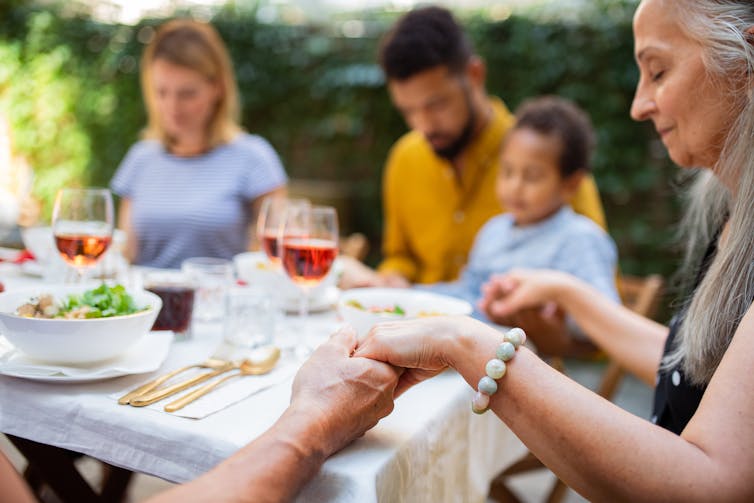 A family holds hands with their eyes shut around a table outdoors.