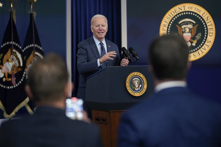 An older man with grey hair stands behind a podium with the U.S. presidential ensign and speaks.