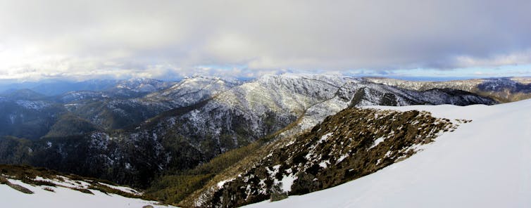 panorama from a mountaintop of other snow-capped, wide mountain ranges below a cloudy sky
