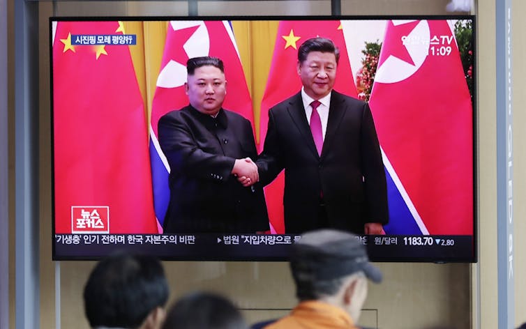 Two men shake hands on a public TV screen as people walk by.