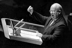 A black and white photo shows a bald, aged man raising his fist while making a speech.