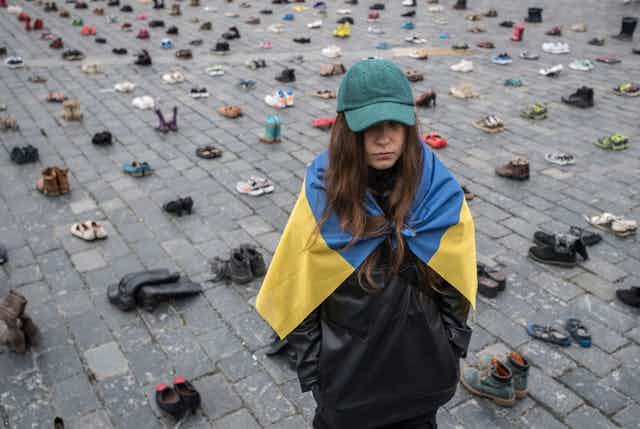 A woman in a teal baseball cap, draped in a Ukrainian flag, stands on pavement covered with neatly arranged pairs of shoes.
