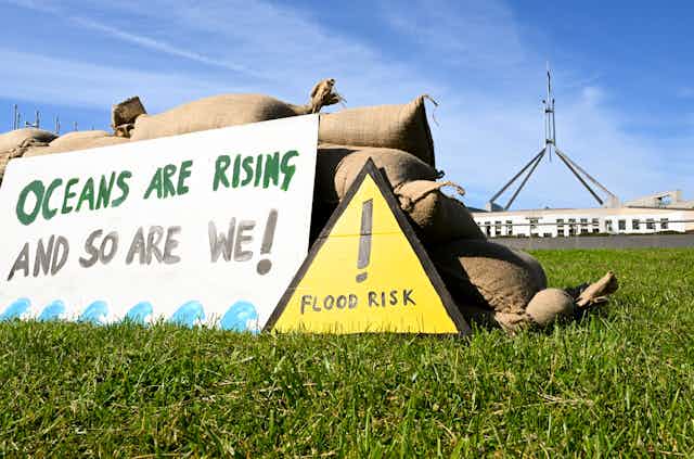 A sign reading: "OCEANS ARE RISING AND SO ARE WE!" and another yellow coloured sign reading: !"FLOOD RISK" on the grass in from of Parliament House