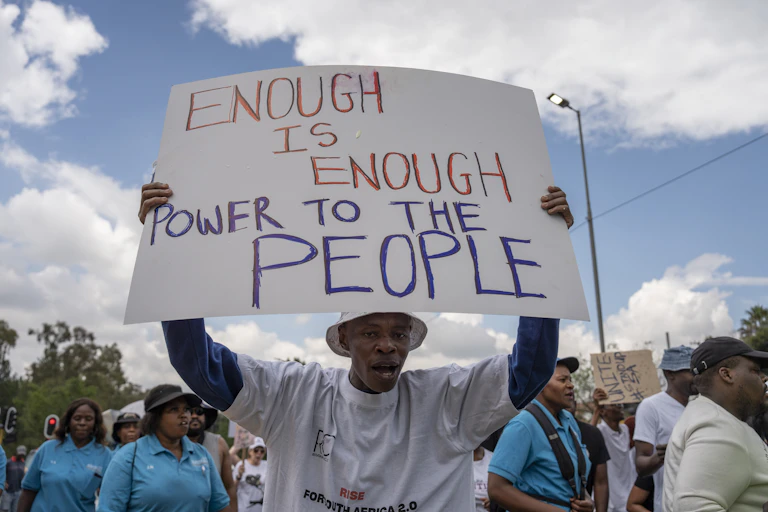 A man in a white shirt and a white bucket hat holds up a placard that reads "Enough is enough, power to the people"