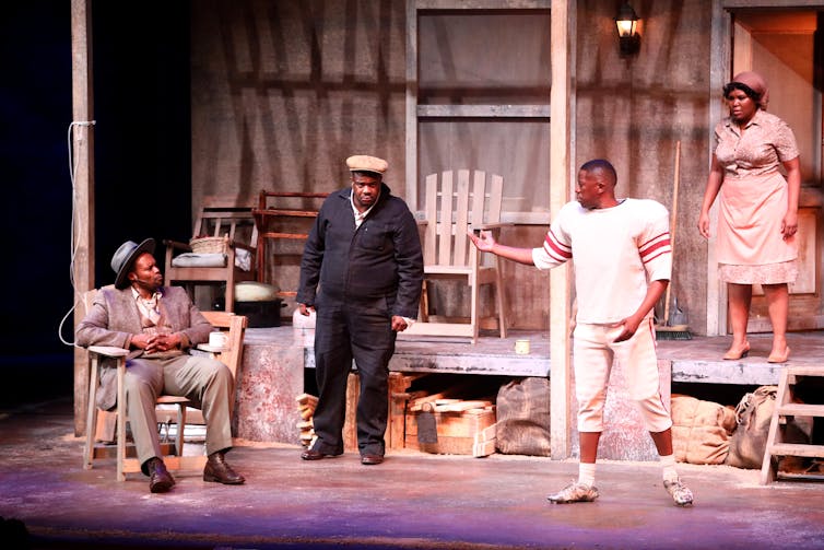 Four people, a woman and three men, are on a porch of a dilapidated house, a young man pointing at an older man in a chair.