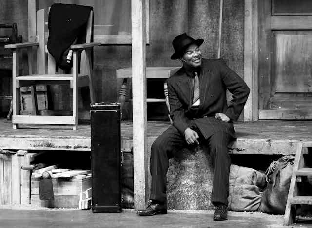 A black and white photo of an actor on stage, a mic down the side of his face. He is dressed smartly in an old-fashioned suit and sits on a porch of a slightly dilapidated house.