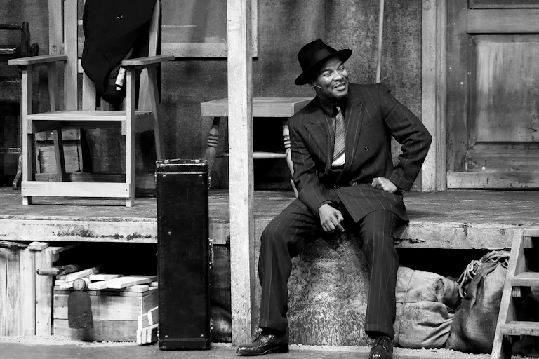 A black and white photo of an actor on stage, a mic down the side of his face. He is dressed smartly in an old-fashioned suit and sits on a porch of a slightly dilapidated house.