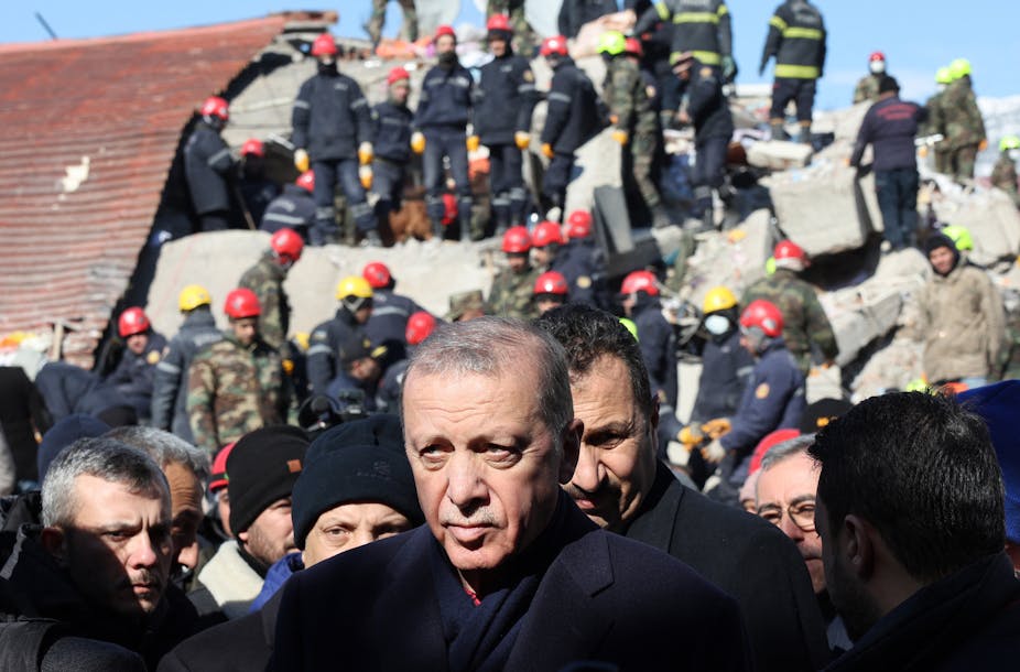 A man in a dark-colored coat stands in front of emergency workers in red and yellow helmets standing on rubble.