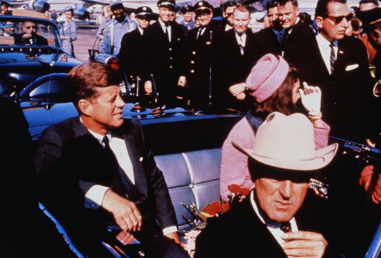 An old colored photo shows a man in a dark suit seated in a car with a lady in a pink suit. Behind them are people in police uniform smiling and watching.