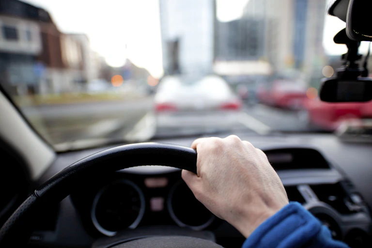 Se ve la mano derecha en el volante de un automóvil y, al frente del parabrisas, otros automóviles.