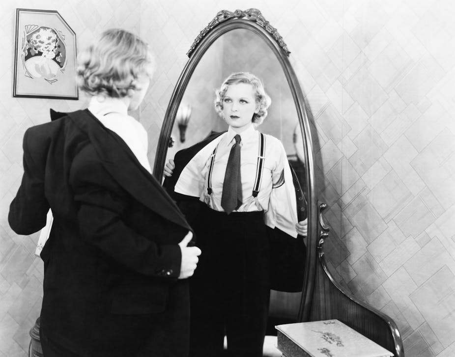 A woman dressing in a suit, with tie and braces, in front of a mirror.