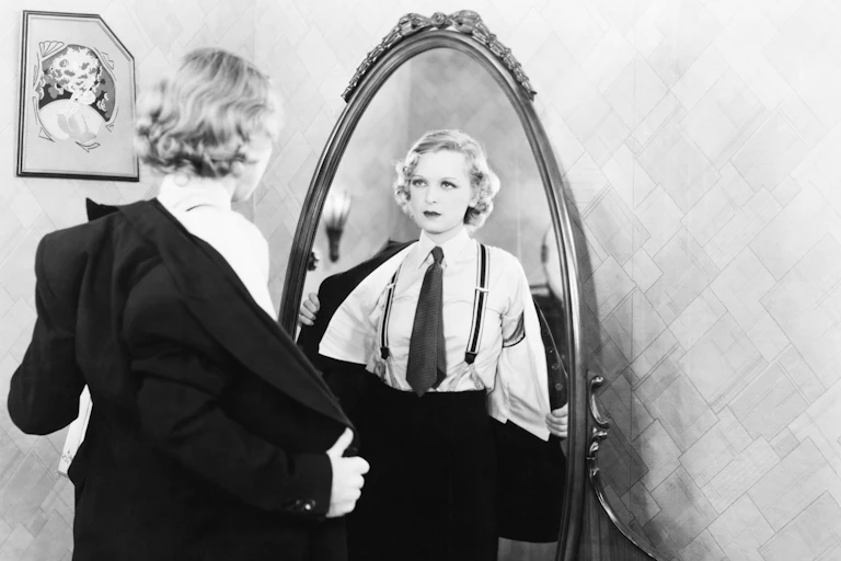 A woman dressing in a suit, with tie and braces, in front of a mirror.