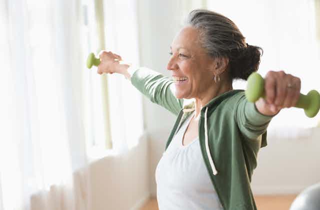 An older woman lifting weights and smiling in her living room.