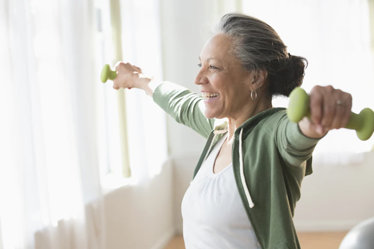 An older woman lifting weights and smiling in her living room.