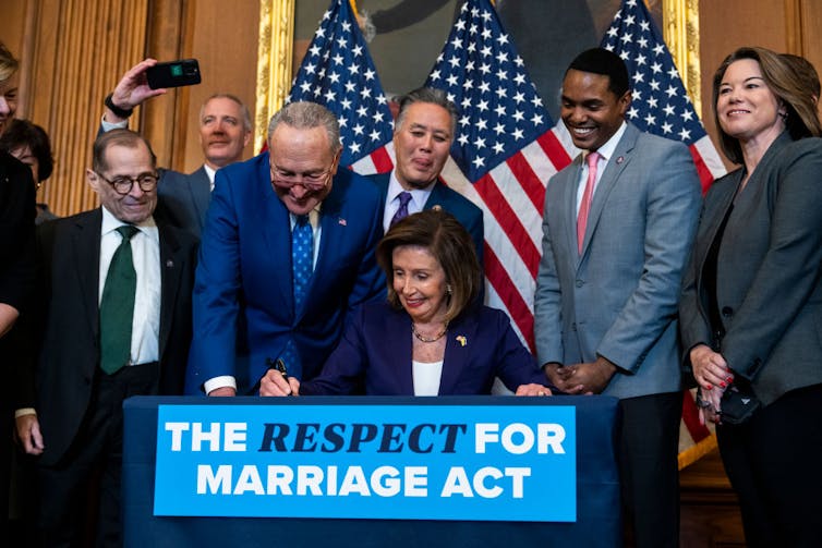 A woman in a blazer signs something on a table with a sign reading 'The Respect for Marriage Act' as people look on.